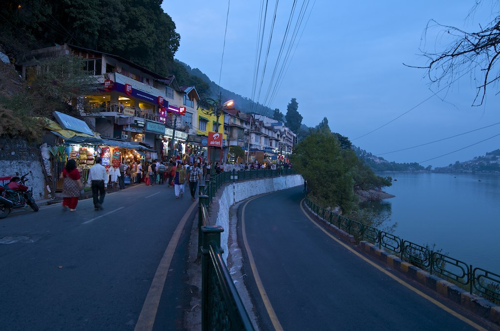 Crowded evening scene at Nainital Mall Road with shops, tourists walking, and Naini Lake visible alongside the road in Uttarakhand