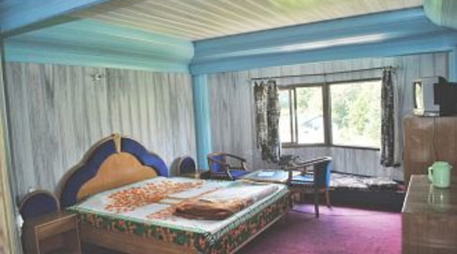 Cozy wooden hotel room with a double bed decorated with flower petals, blue ceiling accents, large window with scenic greenery view, chairs and table near the window, and a TV with a kettle on a side cabinet.