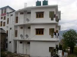 White multi-story guesthouse building with simple balconies, satellite dishes, rooftop water tanks, and surrounding hillside views under a cloudy sky.